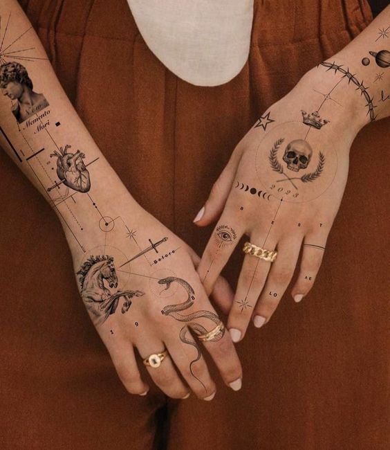 Decorative tattoos on hands featuring skull, horse, eye, and geometric designs, paired with gold rings and brown pants.