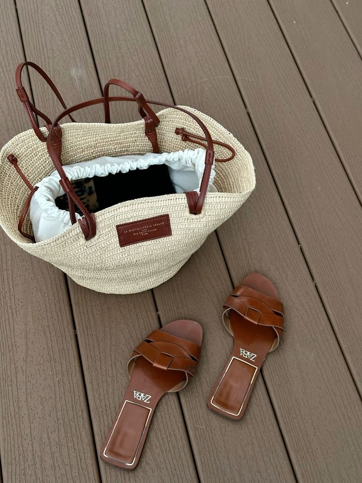 Straw handbag with leather straps and brown sandals on wooden deck flooring.
