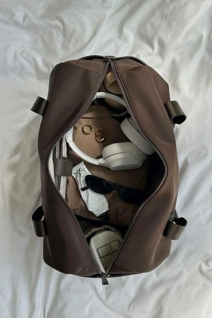 Travel bag with headphones, sunglasses, and essentials on a white sheet for a weekend getaway.