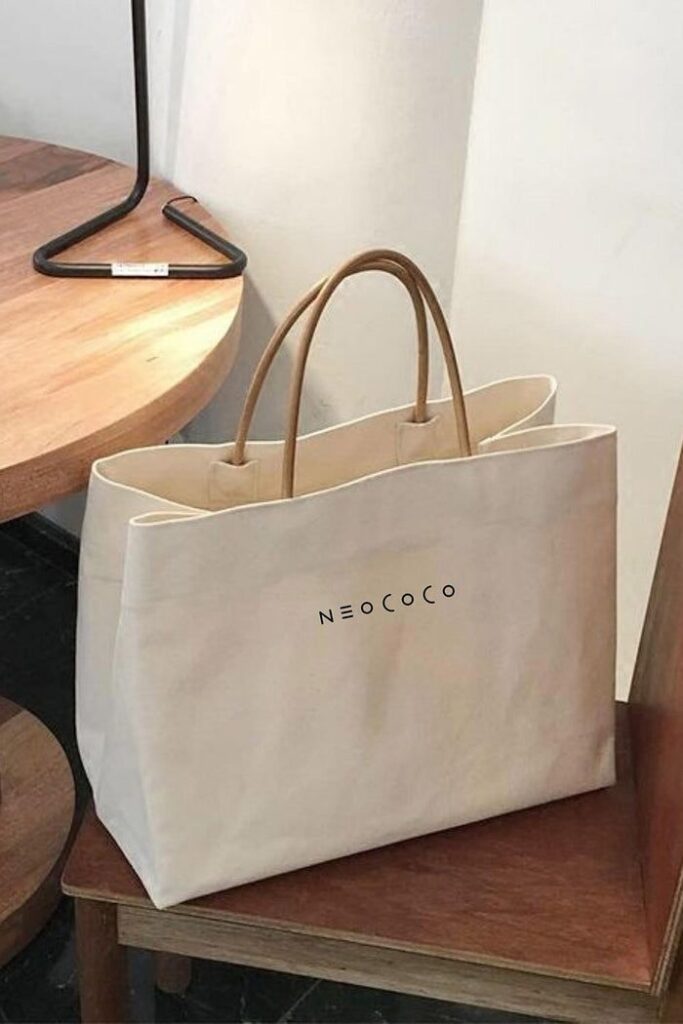 Stylish beige tote bag with NeoCoco logo, placed on a wooden chair near a round table.