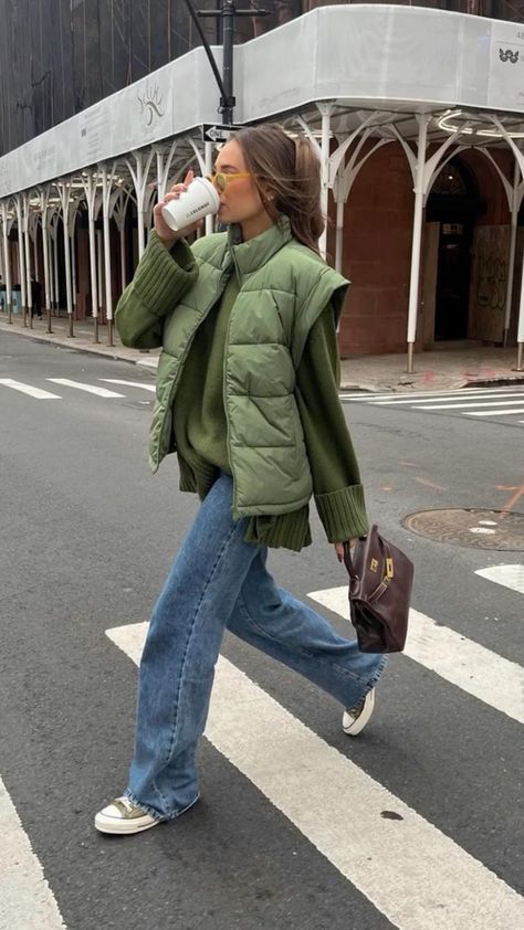 Stylish woman in green vest and jeans crossing street, sipping coffee. Casual urban fashion.