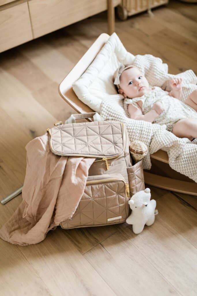 Baby in rocker with beige diaper bag and plush toy on wooden floor, cozy nursery setting.