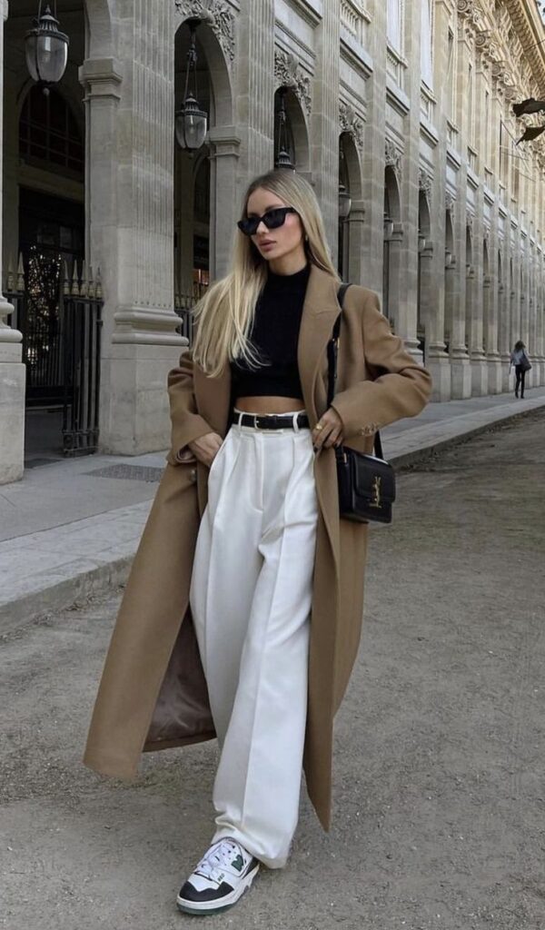 Fashionable woman in long coat and sunglasses walking by elegant building.