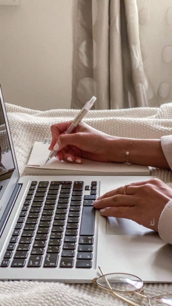 Hands writing in a notebook and typing on a laptop, next to glasses on a cozy blanket.