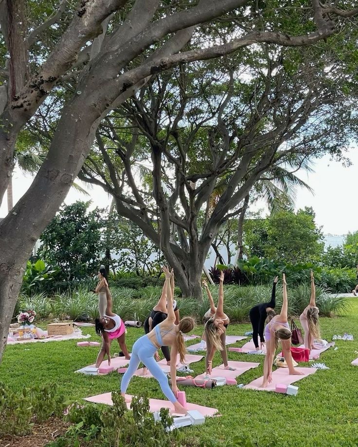 Women practicing yoga outdoors on pink mats, surrounded by lush greenery and large trees.