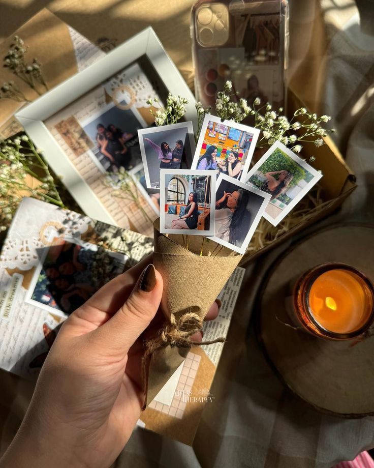 Hand holding a cone of photos with a candle, framed pictures, and scrapbook in the background, evoking nostalgia.