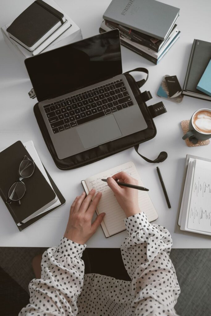Person writing notes at a desk with a laptop, books, glasses, and a coffee cup, creating a productive workspace.