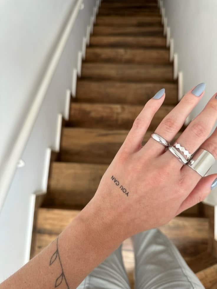 Close-up of a hand with rings and You Can tattoo, resting near a wooden staircase, symbolizing motivation and style.