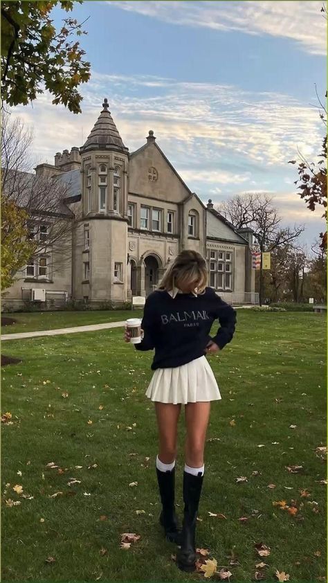 Person in casual autumn outfit holding coffee cup, standing before a historic building with scenic sky and trees.
