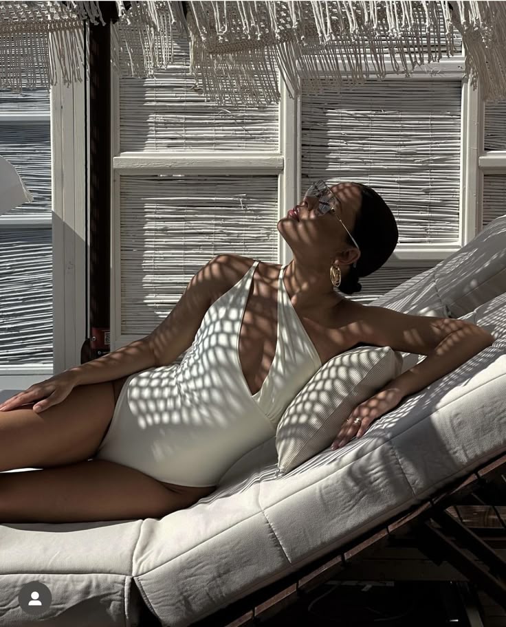 Woman in white swimsuit lounging on a sunbed under dappled light, wearing sunglasses and gold hoop earrings.