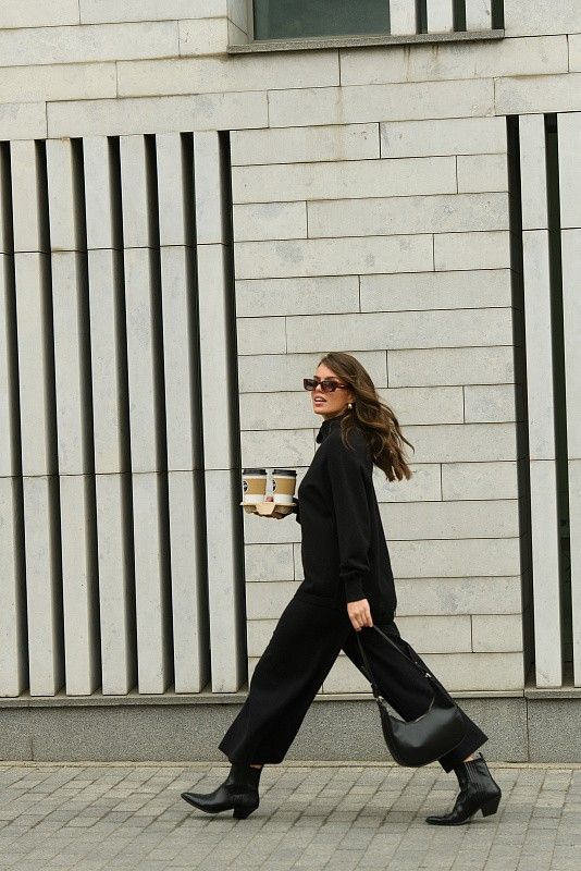 Woman in black outfit walking with coffee cups, smiling against modern building backdrop.