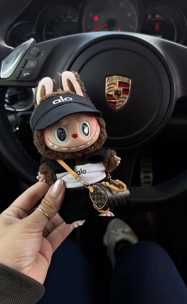Cute toy with Alo visor and designer bag in front of a car steering wheel with a Porsche logo.