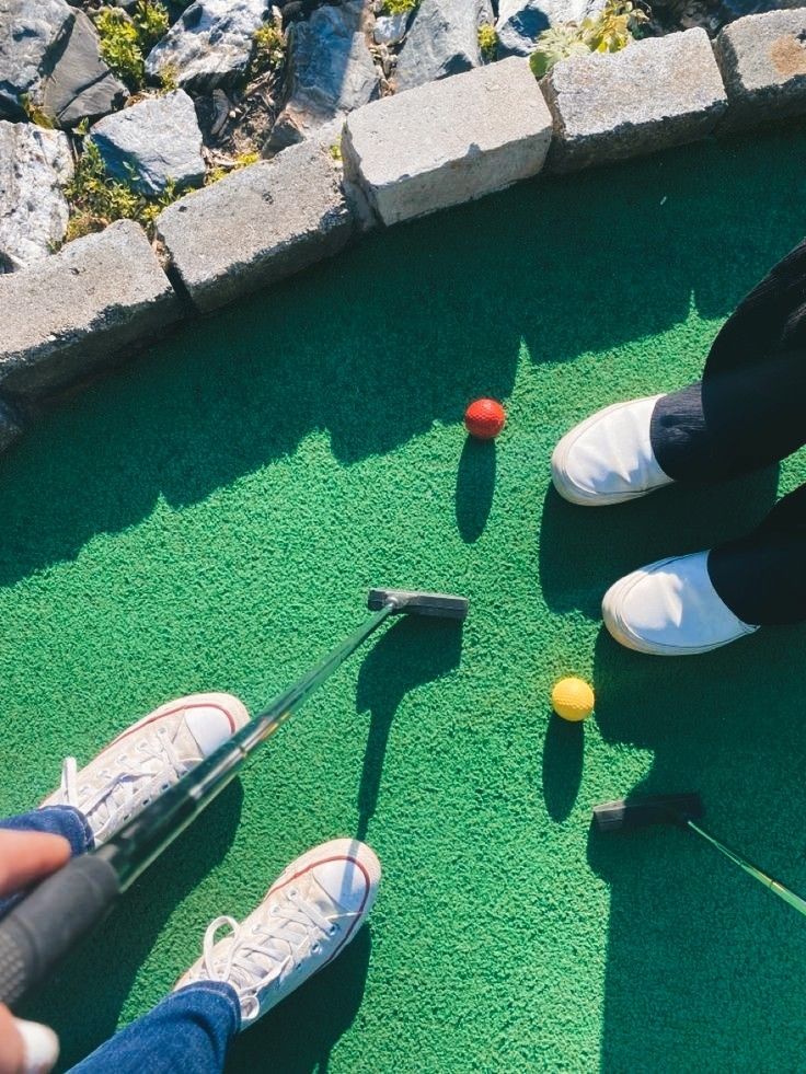 Mini golf game with players holding putters, aiming at red and yellow balls on green turf. Casual shoes visible.