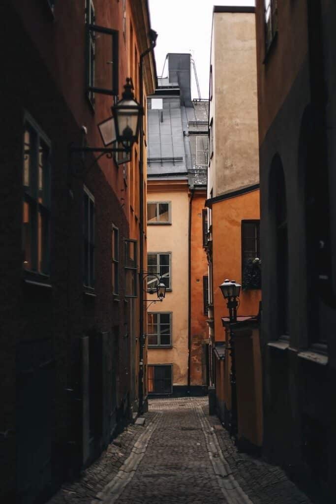 Narrow cobblestone alleyway lined with rustic buildings and street lamps, leading to a distant view of rooftops.