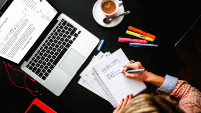 Person writing in notebook beside a laptop and coffee, colorful highlighters on black desk.