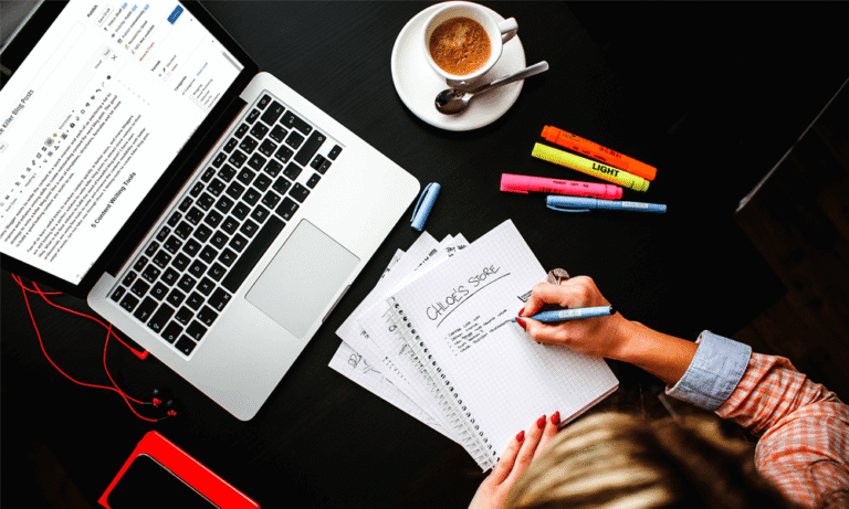 97bc703442fa9a38ed92e3047355fb18486c1219 Person writing in notebook beside a laptop and coffee, colorful highlighters on black desk. | Sky Rye Design Person writing in notebook beside a laptop and coffee, colorful highlighters on black desk.