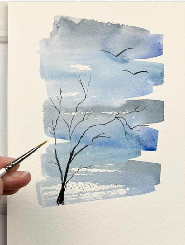 Watercolor painting of bare tree and birds in blue sky, with hand holding brush, evoking serene nature scene.