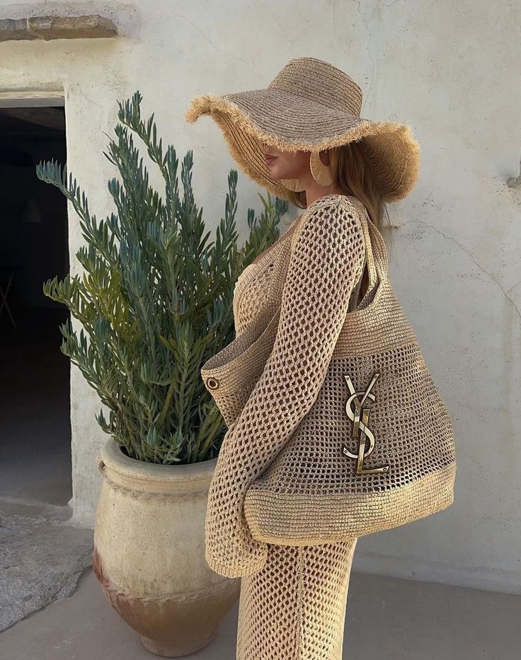 Woman in stylish crochet outfit with oversized straw hat and designer bag, standing by a plant against textured wall.