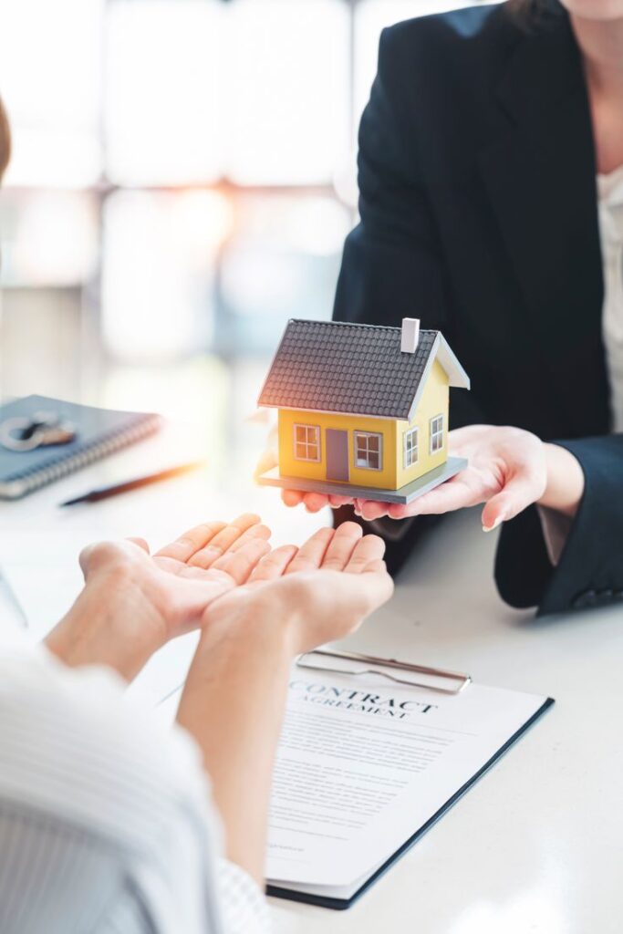 Real estate transaction with hands exchanging a miniature house model over a contract on a desk.