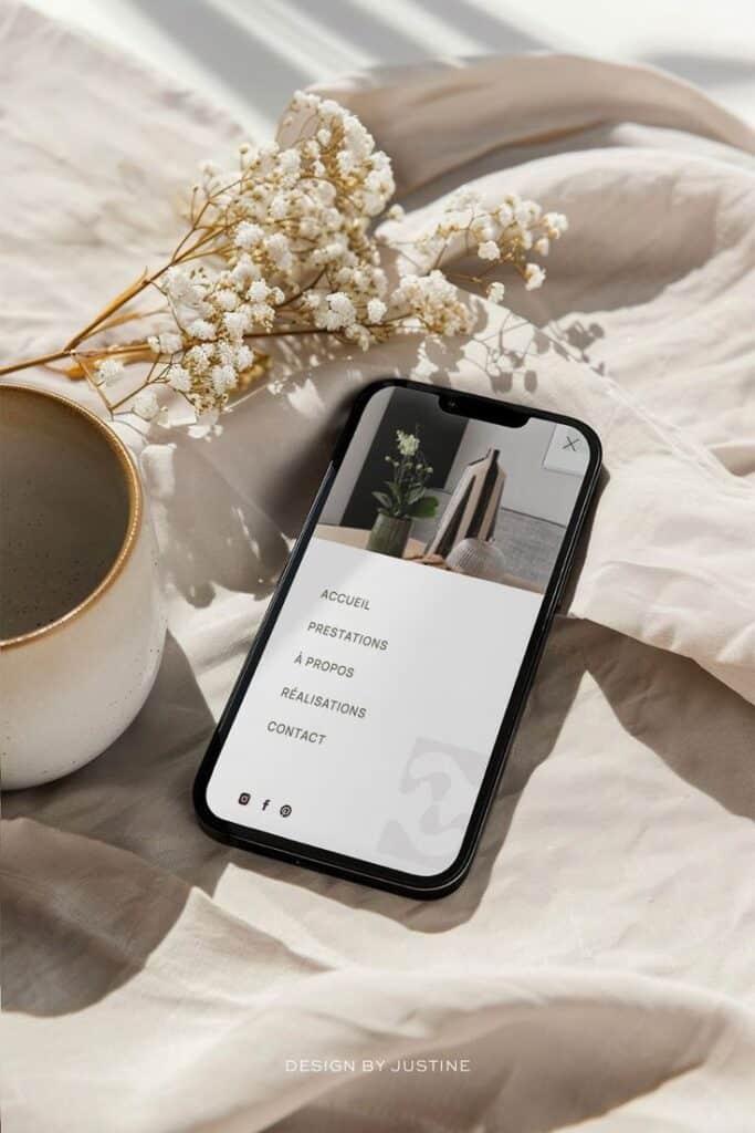 Smartphone with website menu, next to coffee cup and flowers on a beige fabric background.