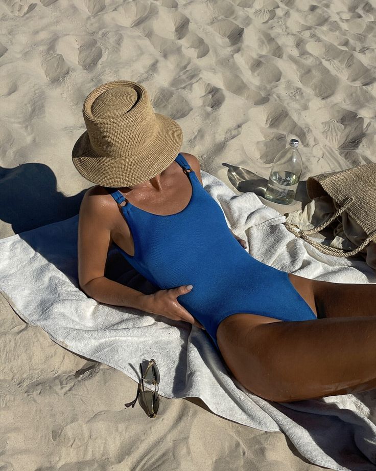 Person in blue swimsuit relaxing on sandy beach with hat, towel, and sunglasses for a summer day.