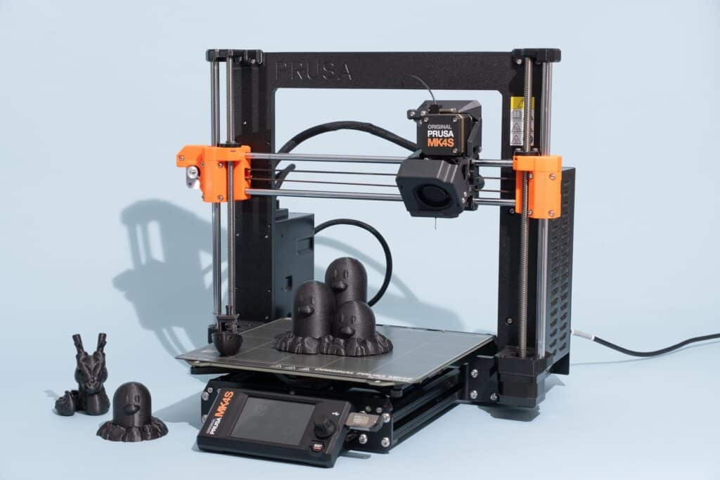 3D printer with black filament models on a blue background, featuring Prusa MK4S components and printed figurines.