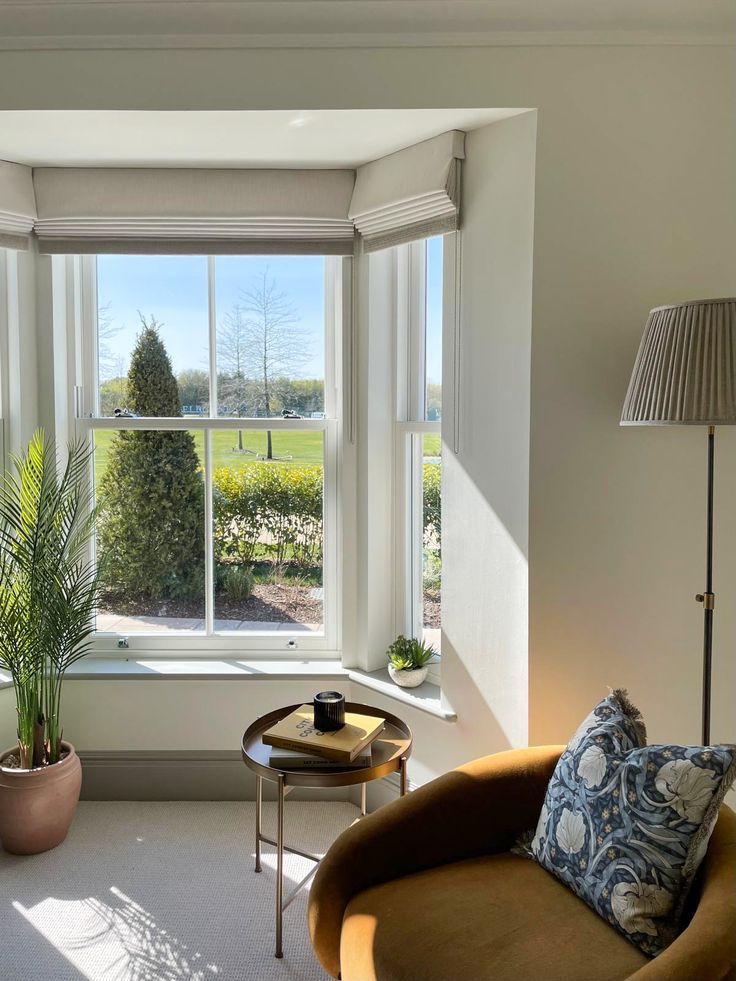 Cozy living room corner with a sunny bay window, potted plant, round table, lamp, and cushioned chair.