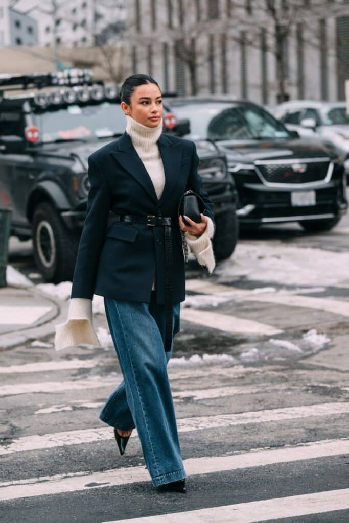 Stylish woman in winter outfit crossing snowy street with confidence, wearing jeans and a belted blazer.