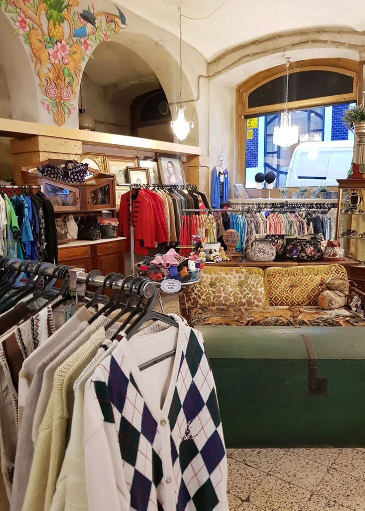 Vintage clothing store interior with racks of colorful garments, cozy sofa, and hanging chandeliers.