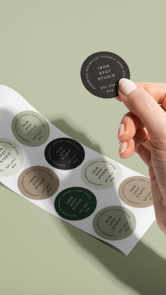 Hand holding a Iron Spot Studio sticker over a sheet of assorted minimalist ceramic home goods labels.