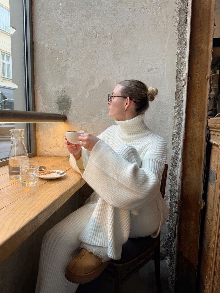Woman in white sweater enjoys coffee by a window in a cozy cafe setting.