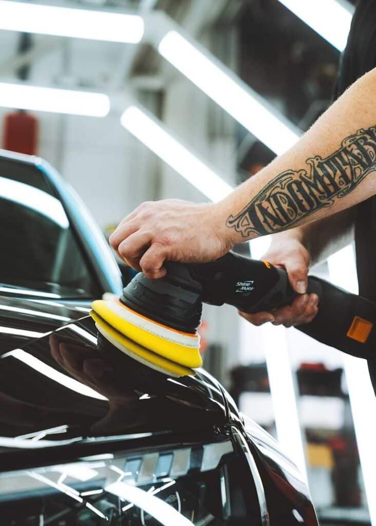 Person polishing a car hood with an electric buffer in a garage setting.