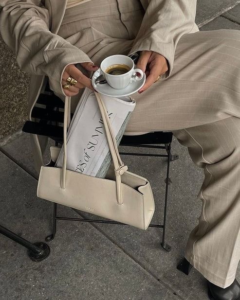 Person in beige outfit holding coffee, white handbag, and a newspaper while sitting outdoors. Elegant and relaxed fashion moment.