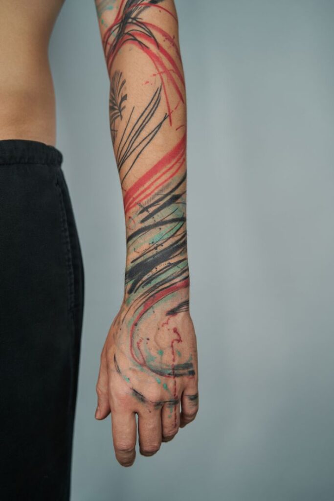 Abstract watercolor tattoo on arm with black, red, and turquoise strokes against a gray background.