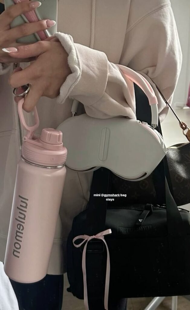Woman holding pink Lululemon water bottle, white headphones, and black Gymshark bag with bow, casual style.