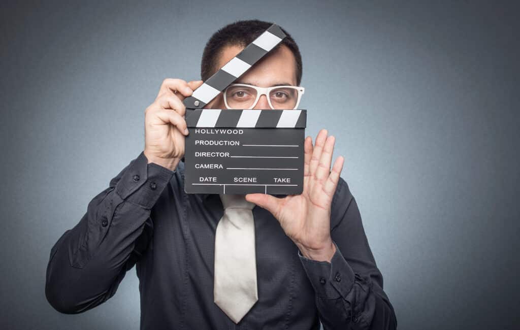 Person with white glasses holding clapperboard on gray background, symbolic of filmmaking or video production.