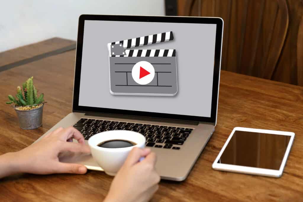 Laptop with video editing icon on screen, person holding coffee at wooden desk with tablet and small cactus.