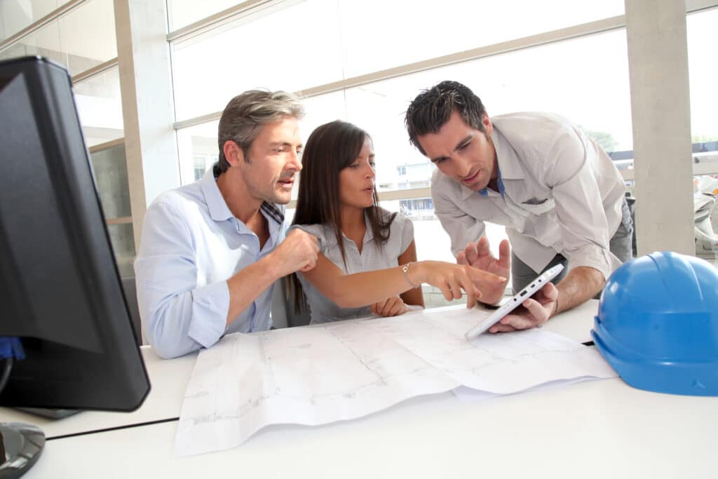 Three architects collaborating on a blueprint with a tablet in a modern office setting.