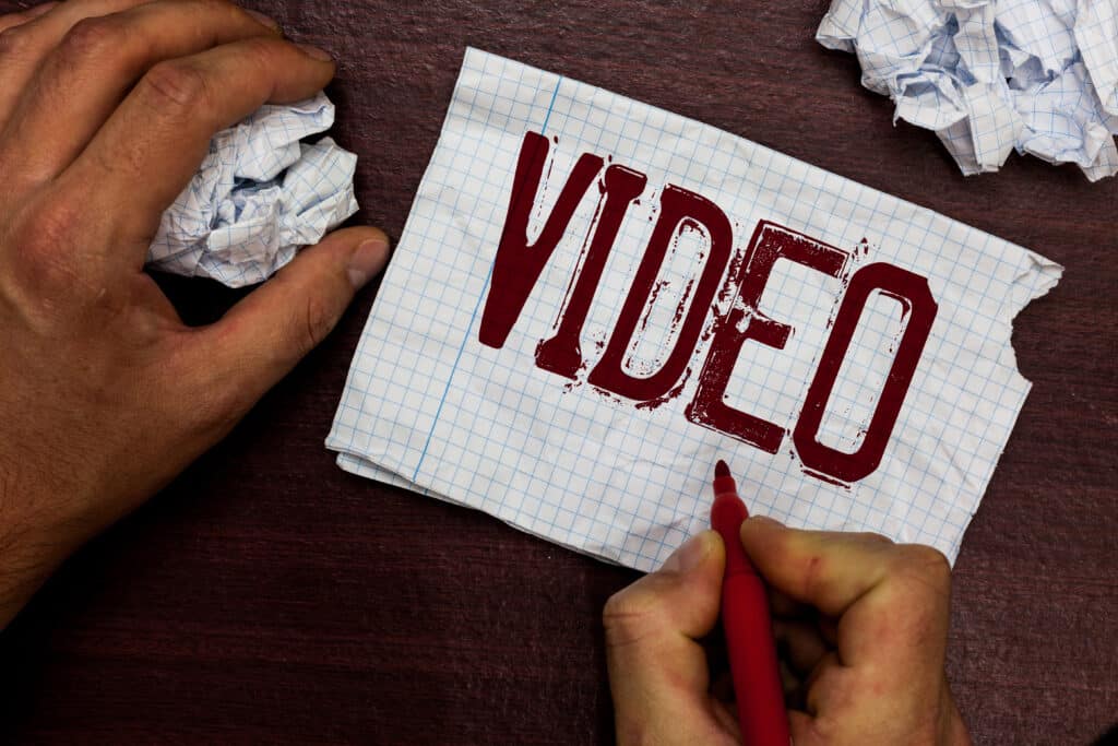 Person writing VIDEO on graph paper with red pen, surrounded by crumpled sheets on a wooden desk.