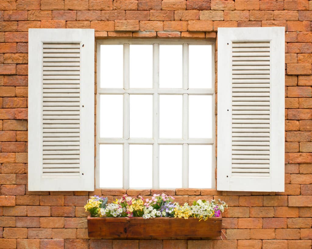 White window with shutters against red brick wall, featuring a colorful flower box.