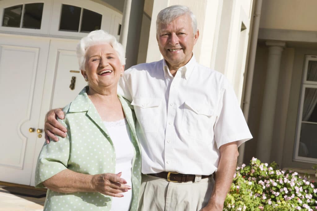 Happy elderly couple smiling in front of their home with flowers in the background.