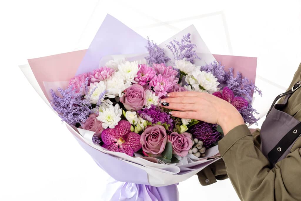 Hand arranging a bouquet of purple, pink, and white flowers wrapped in pastel paper.