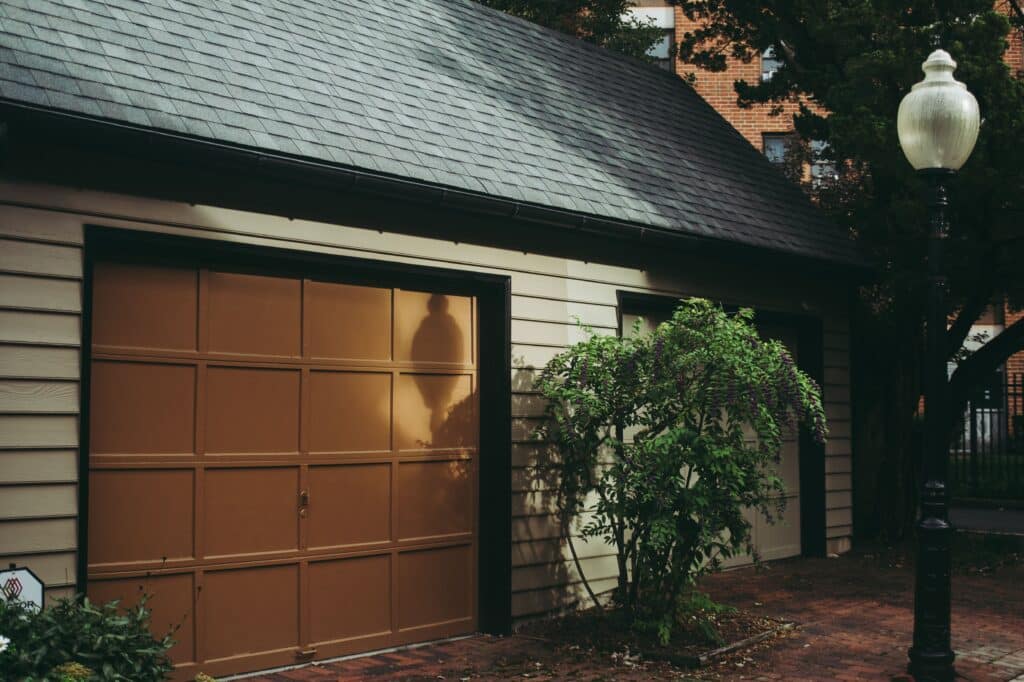 Garage with brown door and shadow of a streetlamp, adjacent green bush with flowers, brick path in front.