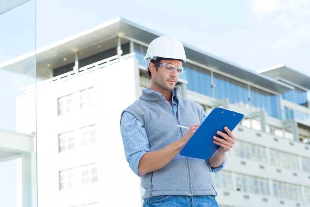 Construction worker in hard hat inspecting site with clipboard, standing in front of modern building.