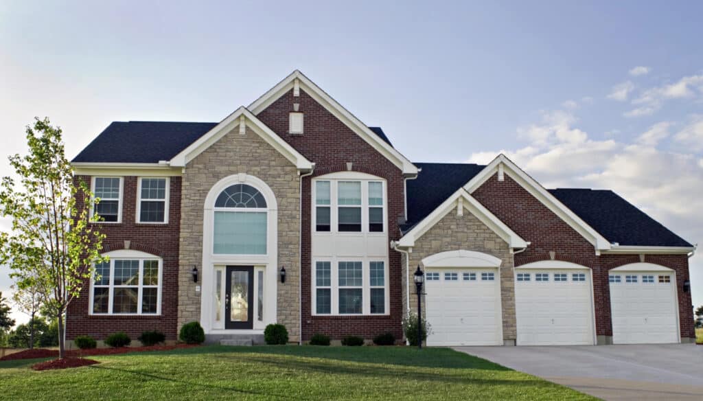 Elegant brick and stone suburban house with three garage doors and lush green lawn under clear sky.