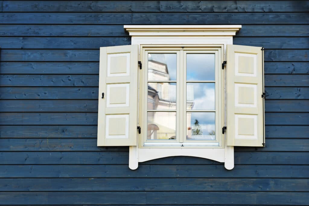 White-framed window with shutters on blue wooden wall reflecting sky and clouds.