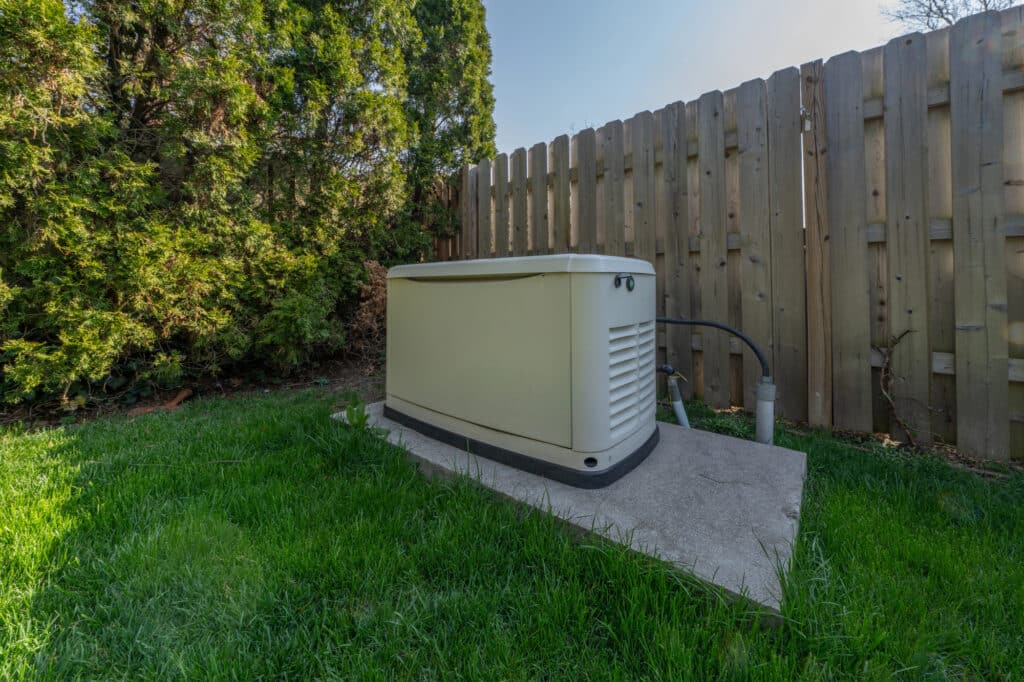 Depositphotos_813021384_L Outdoor standby generator on a concrete slab near a wooden fence surrounded by green grass and shrubs. | Sky Rye Design Outdoor standby generator on a concrete slab near a wooden fence surrounded by green grass and shrubs.
