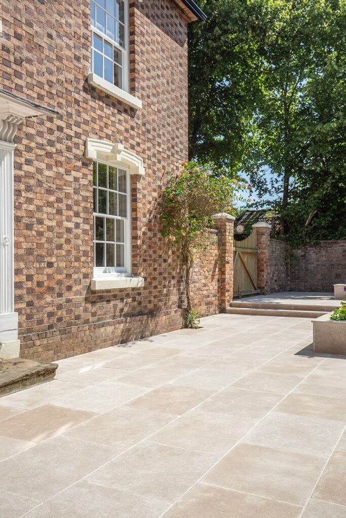 Brick house exterior with spacious stone patio, large windows, and trees in the background, basking in sunlight.