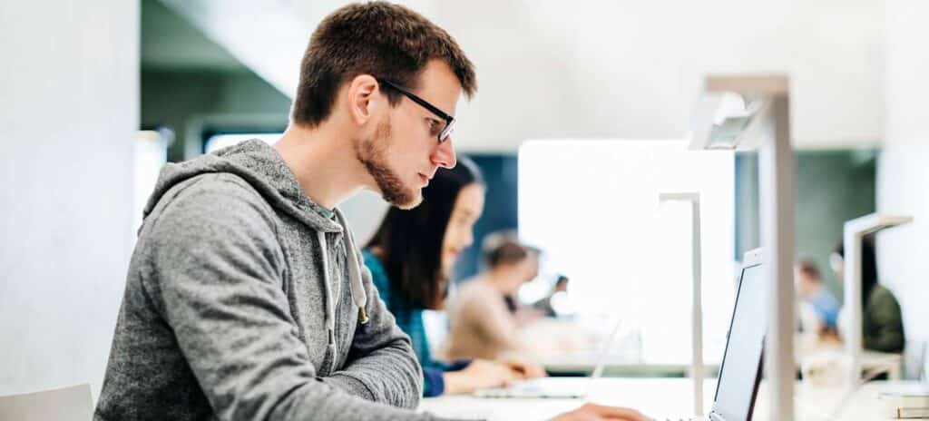 Focused man in glasses and gray hoodie working on laptop in busy office or library setting.