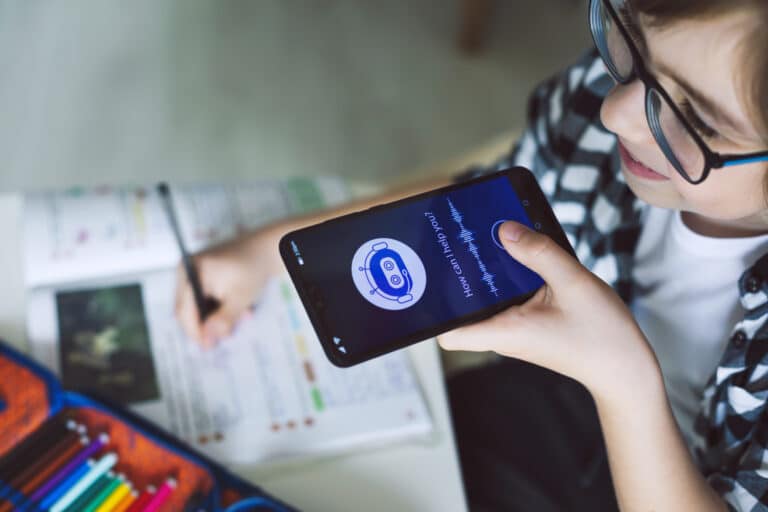 Child using smartphone voice assistant for homework with colorful pencils and book.